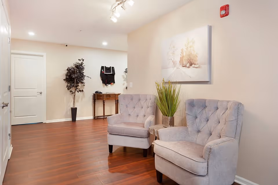 A cozy seating area in a memory care facility with two gray tufted armchairs, a small side table with a green plant, a wooden console table against the wall, a potted artificial tree, and a framed picture of a snowy landscape on the beige wall. The floor is a polished wood laminate and the ceiling has recessed and track lighting.