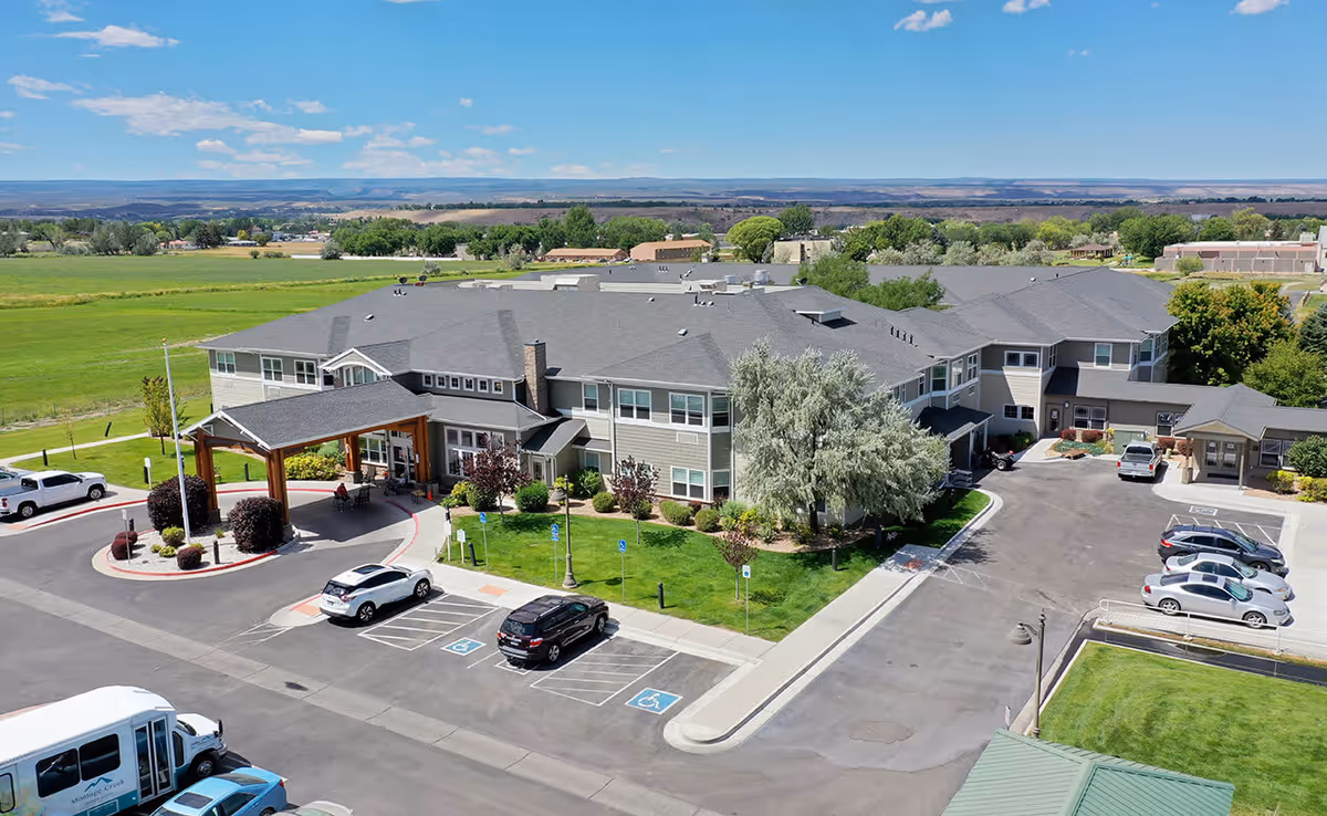 Aerial view of Montage Creek a Generations Community senior living facility showing a large two-story building with a covered entrance, surrounded by parking spaces including handicapped spots, green lawns, and trees under a blue sky with scattered clouds.