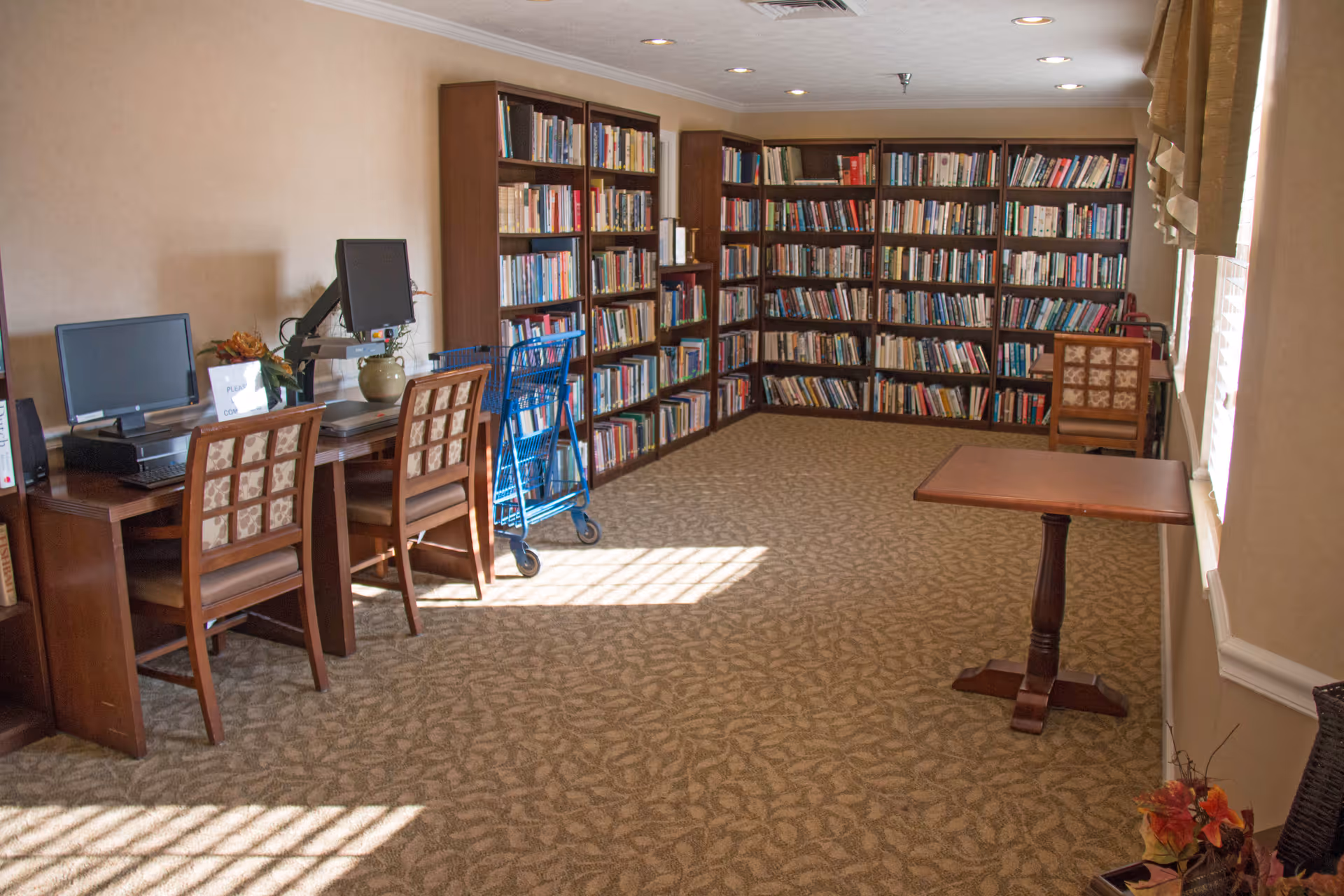 A quiet library room with carpeted floor, several wooden bookshelves filled with books along the walls, two computer workstations with chairs on the left side, a blue book cart, and a small wooden table near a window with blinds letting in sunlight.