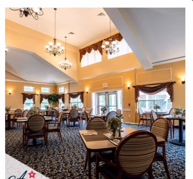 Spacious, well-lit assisted living dining room with multiple tables and chairs, chandeliers, and large windows.