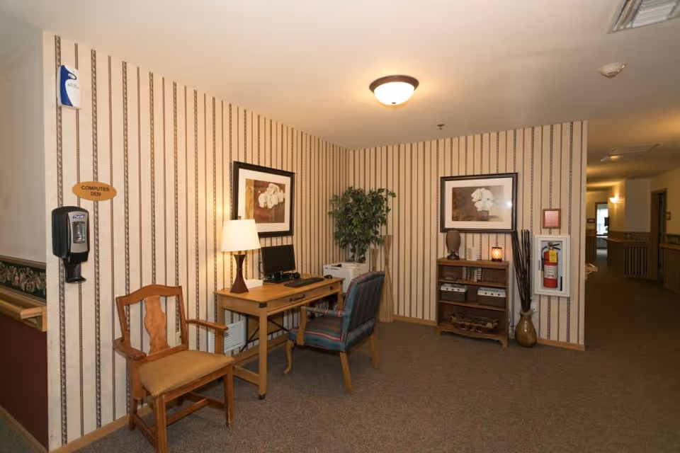 A small computer den area in a senior living facility with striped wallpaper and carpeted floor. The space includes a wooden desk with a computer, a lamp, and a striped upholstered chair. Next to the desk is a wooden chair with armrests. On the wall above the desk are two framed floral pictures. A small wooden bookshelf holds decorative items and books. A fire extinguisher is mounted on the wall nearby, and a potted plant is placed in the corner. A hand sanitizer dispenser is attached to the wall on the left side.