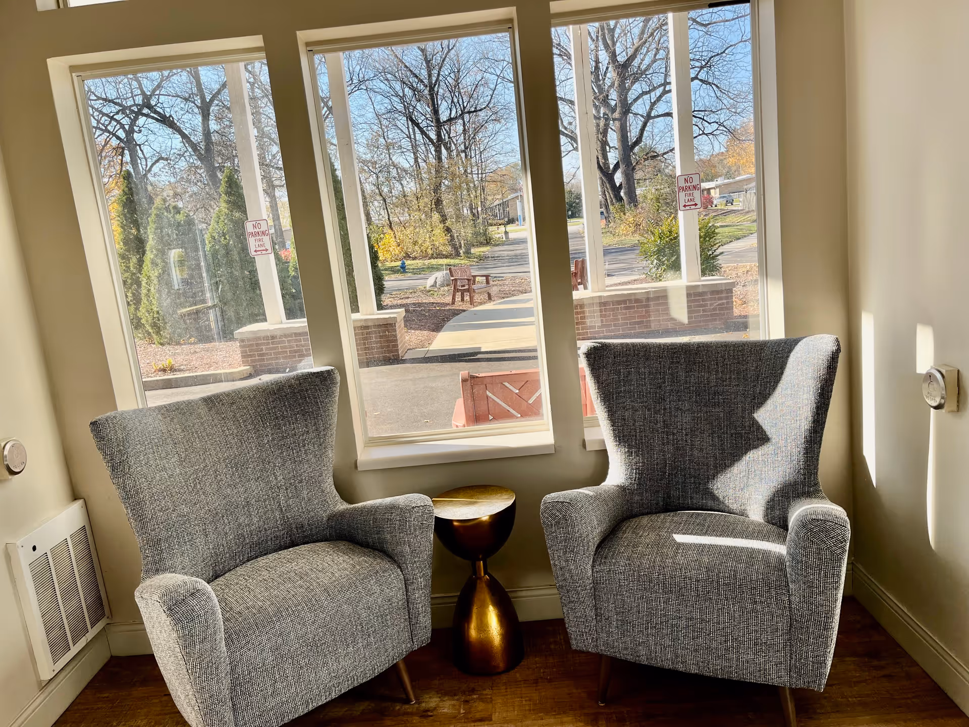 Two gray upholstered armchairs with wooden legs positioned side by side in front of three large windows showing an outdoor view with trees and a walkway. A small, round, gold-colored side table is placed between the chairs. The room has light-colored walls and wooden flooring.