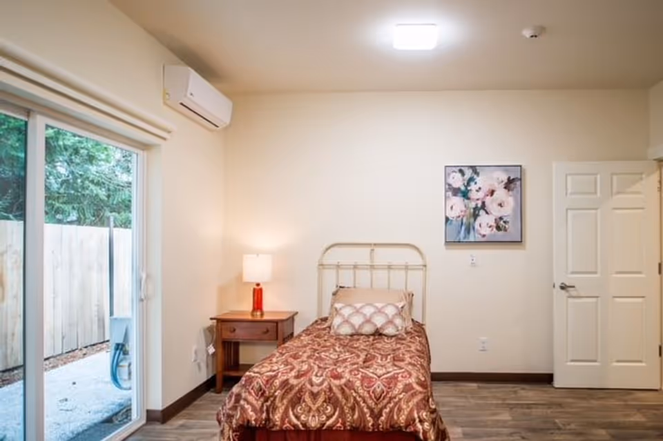 A small bedroom with a single bed covered in a patterned brown and red bedspread, a decorative pillow, a wooden nightstand with a red lamp, a floral painting on the wall, a white door, and a sliding glass door leading outside to a fenced area.