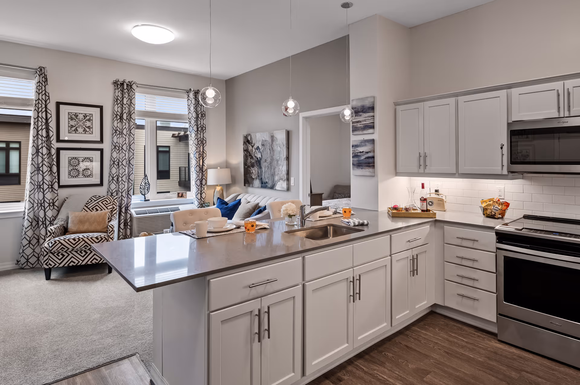 Modern kitchen with white cabinets, stainless steel appliances, and a gray countertop island with a sink. The kitchen opens into a living area with a patterned armchair, a sofa with blue and beige pillows, and large windows with patterned curtains letting in natural light. There are pendant lights hanging above the kitchen island and artwork on the walls.