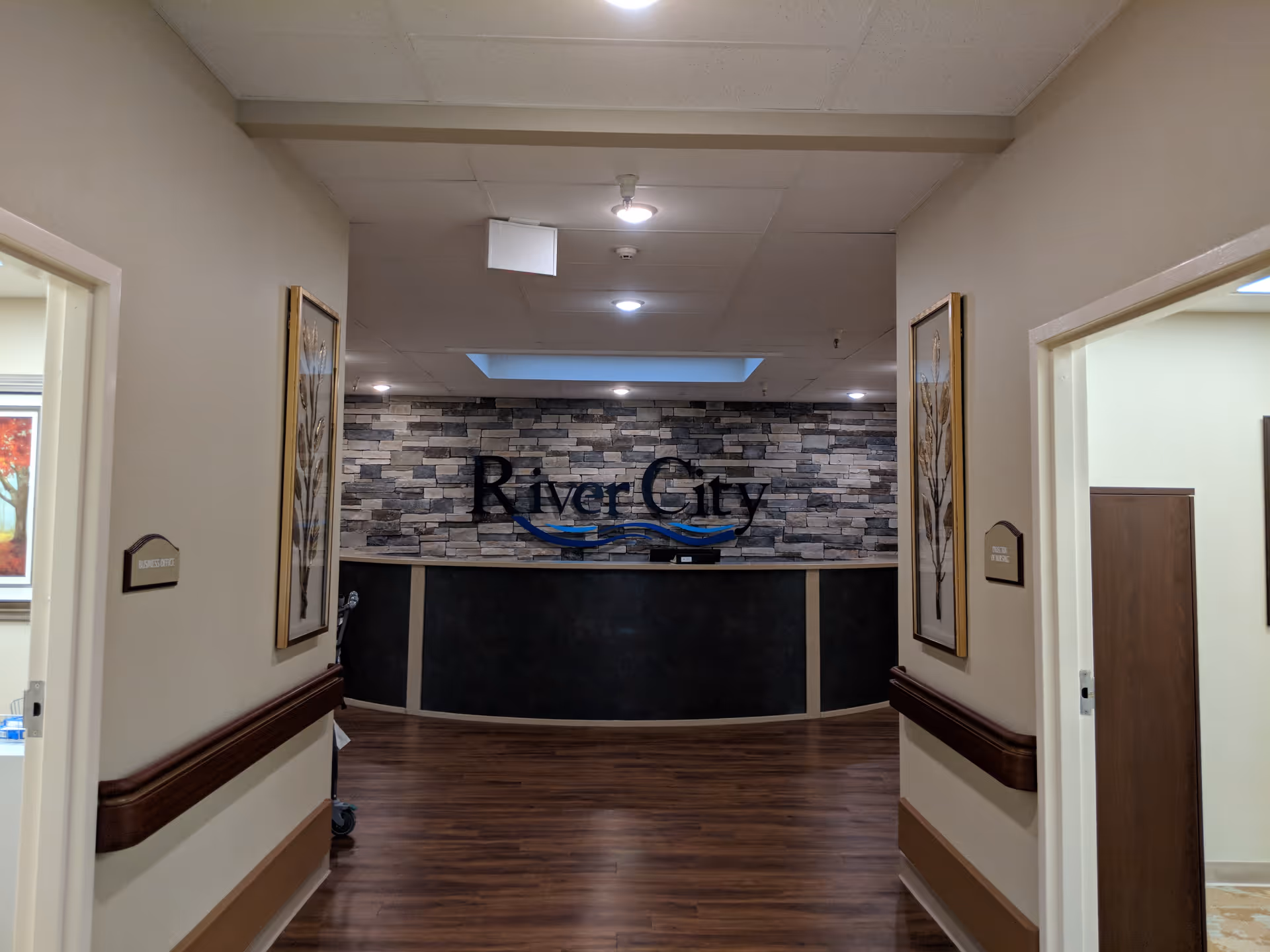 View down a hallway with wood flooring leading to a reception desk with a stone brick wall behind it. The wall has the words 'River City' displayed prominently. The hallway walls have framed artwork and handrails on both sides, with doorways on the left and right.