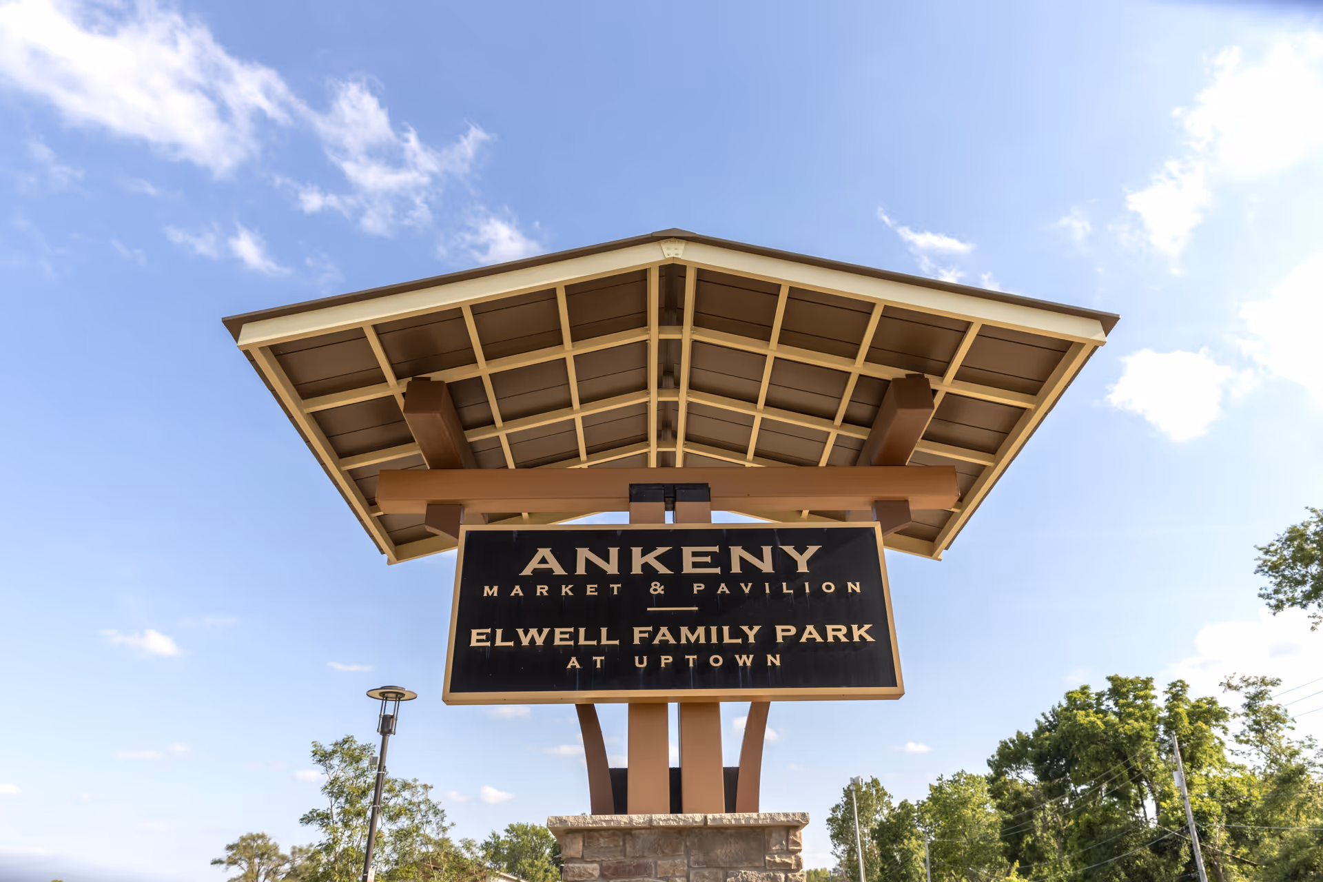 Wooden pavilion sign for Ankeny Market & Pavilion / Elwell Family Park beneath a large shelter structure against a blue sky.