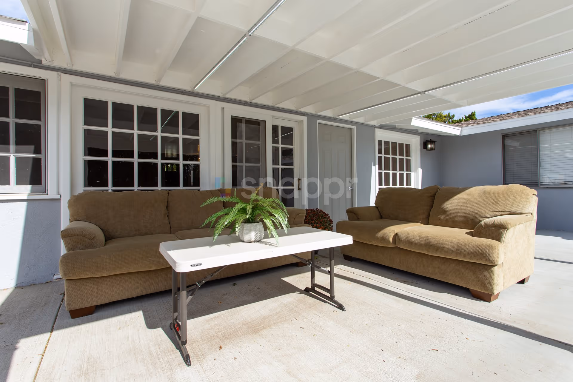 Outdoor covered patio area with two brown sofas facing each other and a white folding table with a potted green plant on top. The background shows windows and doors of a building under the covered roof.