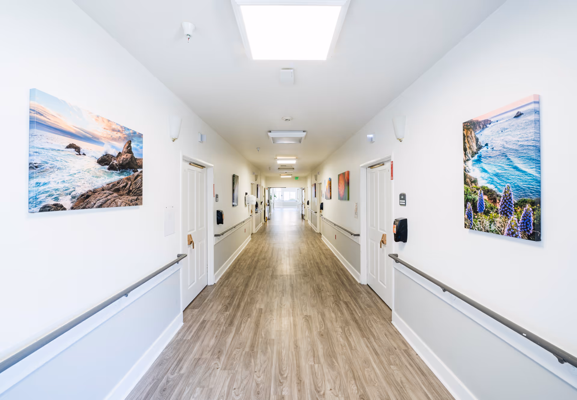 A long, bright hallway with wood-patterned flooring and white walls. The hallway has handrails on both sides and several closed white doors. Colorful landscape paintings featuring ocean scenes are hung on the walls. Ceiling lights illuminate the corridor, and there is a window at the far end letting in natural light.