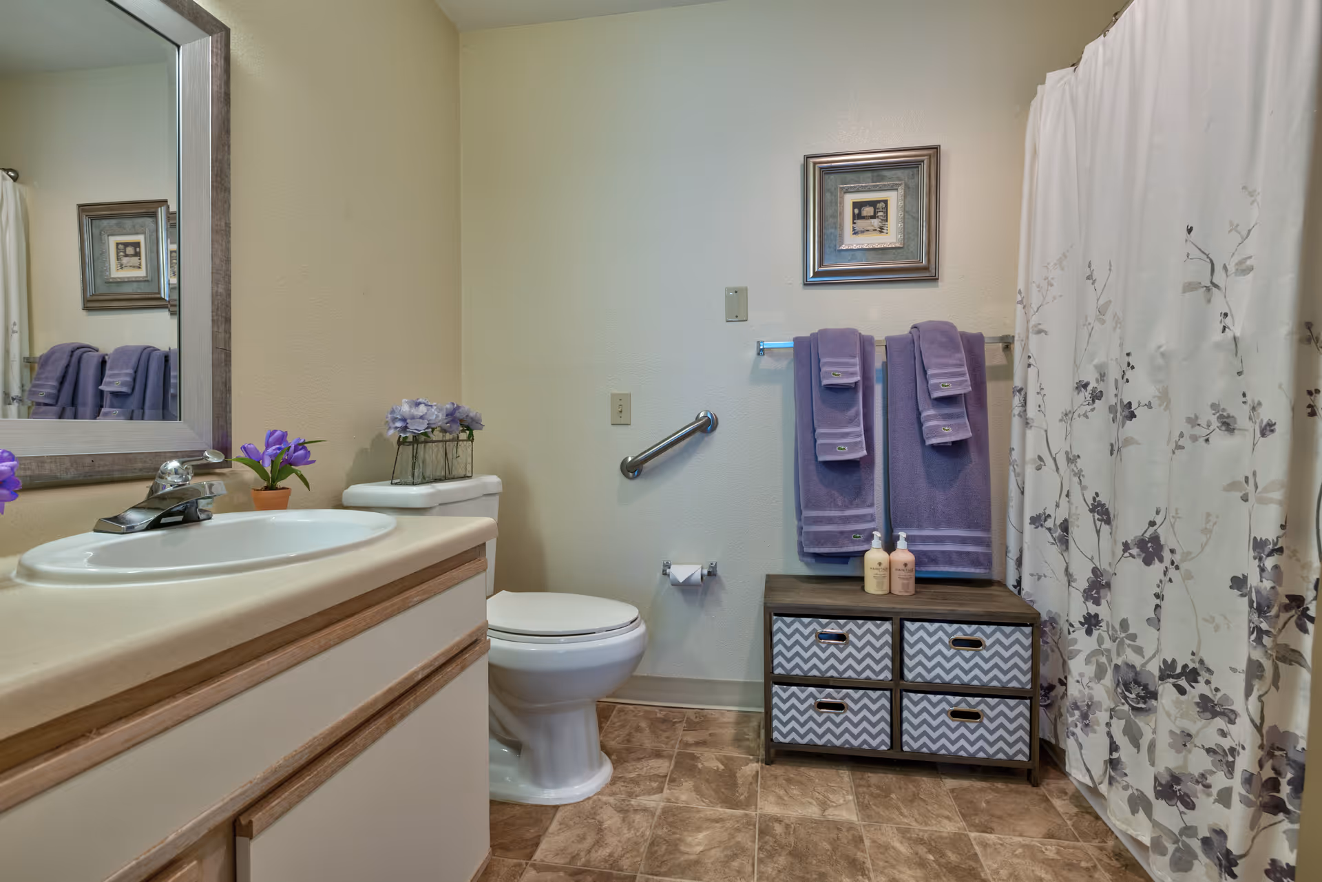 A bathroom with a white sink and countertop on the left, a toilet in the center, and a shower with a floral curtain on the right. There are purple towels hanging on a rack above a small storage unit with four drawers. A framed picture hangs on the wall above the towels, and there are small potted plants on the sink and toilet tank.