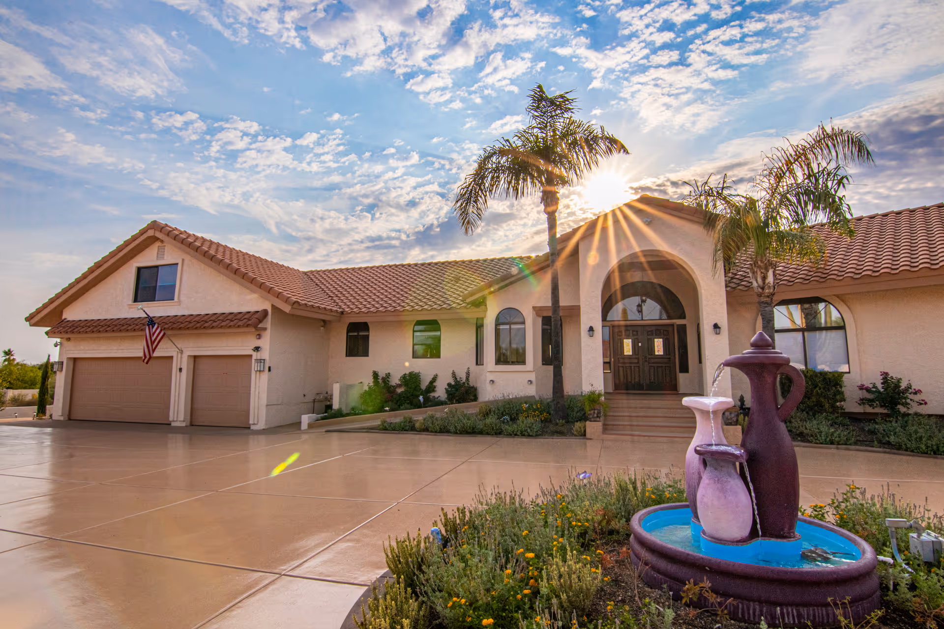 Exterior view of a single-story assisted living facility with a tiled roof, an American flag near the garage, palm trees, and a decorative water fountain in the foreground under a partly cloudy sky with the sun shining.
