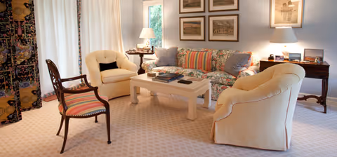 A cozy living room with a floral patterned sofa adorned with striped and solid pillows, two cream-colored armchairs, and a wooden chair with striped upholstery. A white coffee table with books sits in the center on a patterned carpet. The room is softly lit by two table lamps on side tables, with framed artwork on the wall and a window with white curtains.