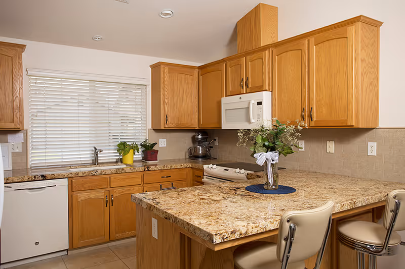 A kitchen with wooden cabinets, granite countertops, a white microwave above a white stove, a dishwasher, and a sink under a window with white blinds. There are two beige bar stools at the counter, and a vase with flowers on the countertop.