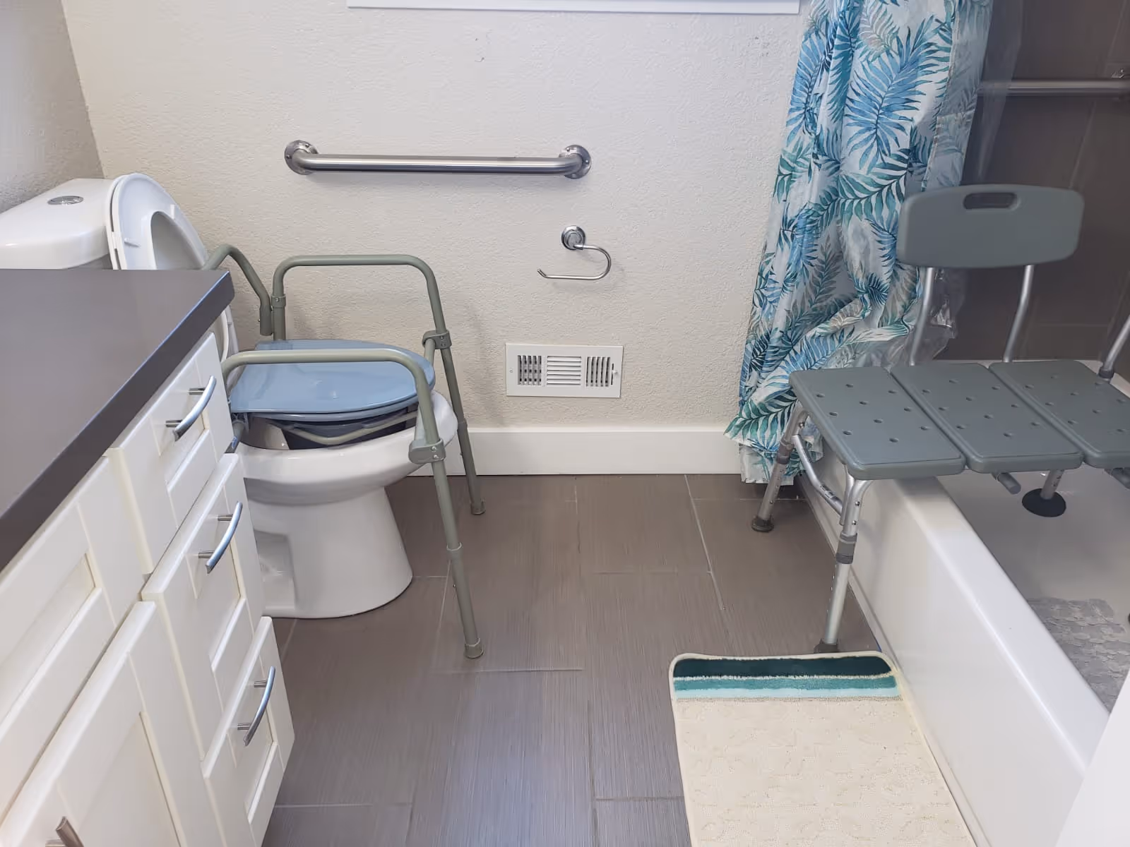 A bathroom with a white toilet equipped with a gray commode chair, a white vanity with drawers on the left, a grab bar on the wall above the toilet paper holder, a bathtub with a green and white leaf-patterned shower curtain, and a gray shower chair placed over the bathtub. A white and green bath mat is on the floor in front of the tub.