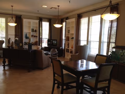 A well-lit living room area with large windows covered by brown curtains, a wooden dining table with four chairs, a sofa, a TV on a stand, built-in white bookshelves filled with decorative items, and a fish tank on a wooden cabinet.