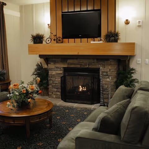 Cozy living room with a stone fireplace featuring a lit fire, a wooden mantel with decorative plants and a small bicycle ornament, a flat-screen TV mounted above the mantel, a round wooden coffee table with a floral arrangement, and a green upholstered sofa.