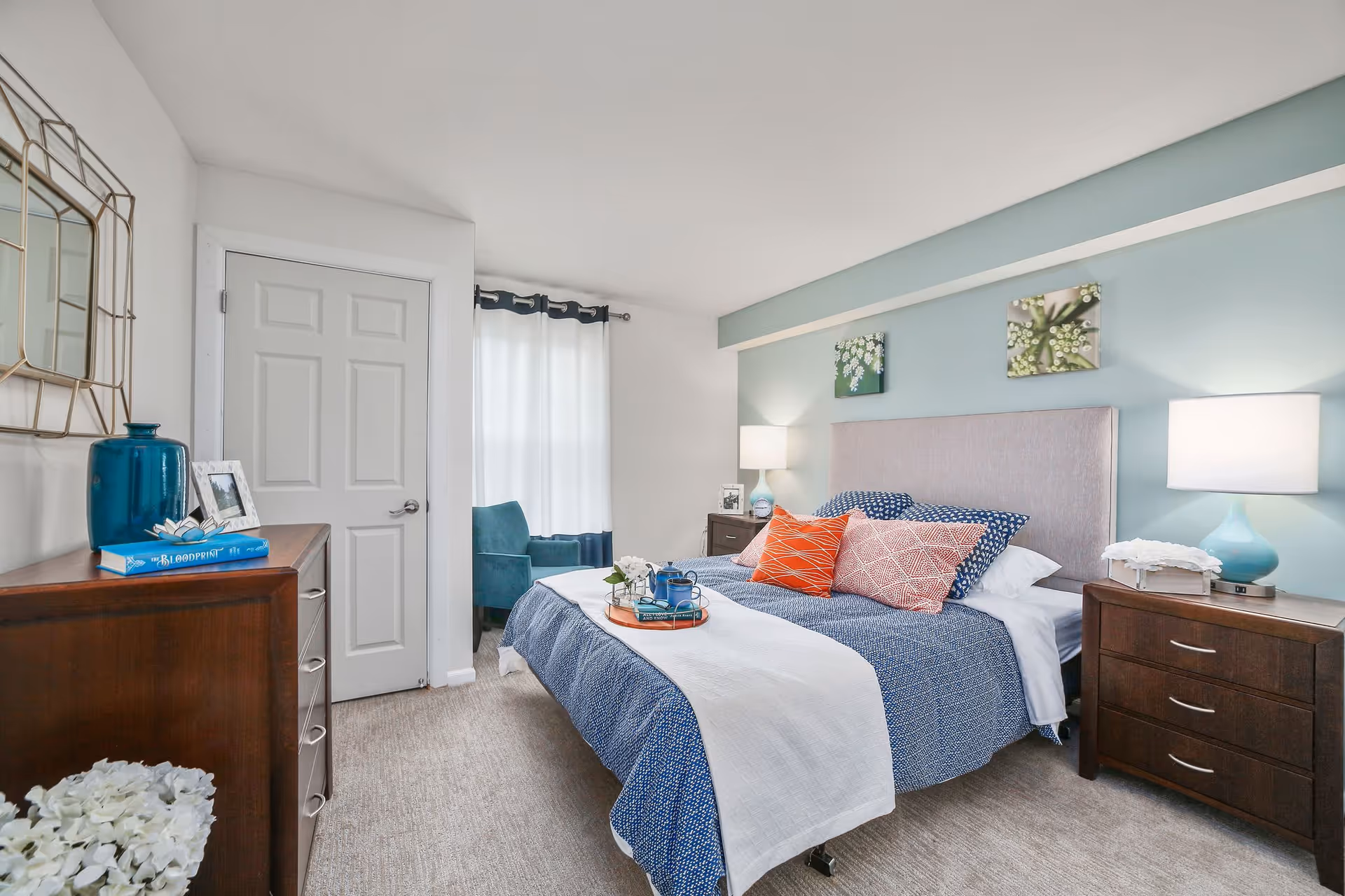A bright and cozy bedroom with a large bed featuring blue and white bedding and multiple colorful pillows. There are two wooden nightstands with lamps on either side of the bed. A wooden dresser with a blue vase, a framed photo, and a book is on the left side of the room. A window with white curtains and a blue armchair are visible in the background. The walls are painted white and light blue, with two floral paintings above the bed.