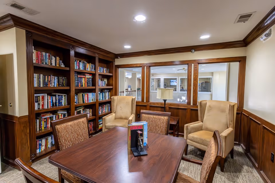 A cozy library or reading room with wooden bookshelves filled with books, a wooden table surrounded by four chairs, and two beige upholstered armchairs. The room has wood paneling on the lower walls and a window looking into another interior space. A table lamp is visible on a small side table near the window.
