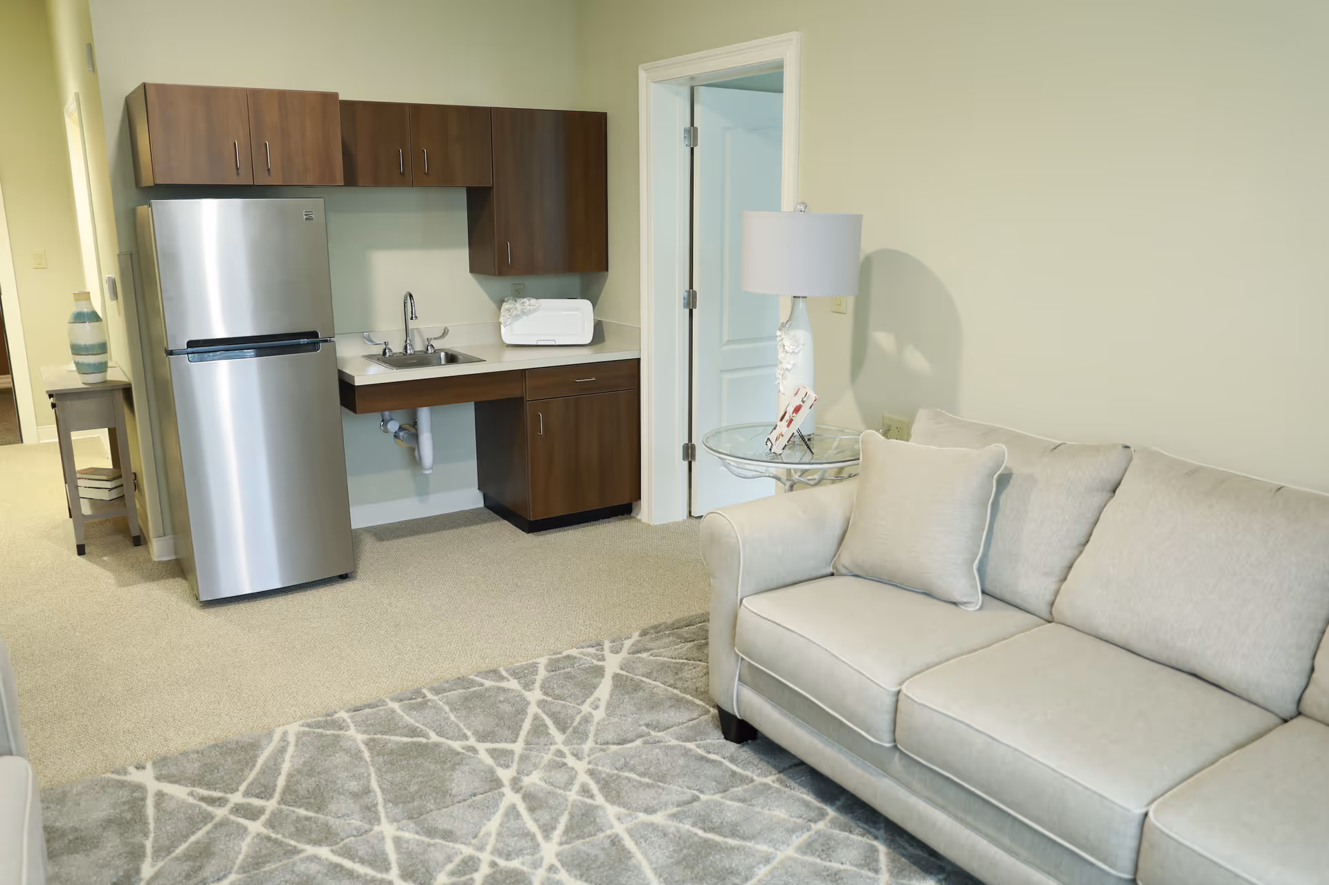 A bright interior living area with a small kitchenette featuring a stainless refrigerator and sink, a beige sofa, side table with lamp, and patterned rug.
