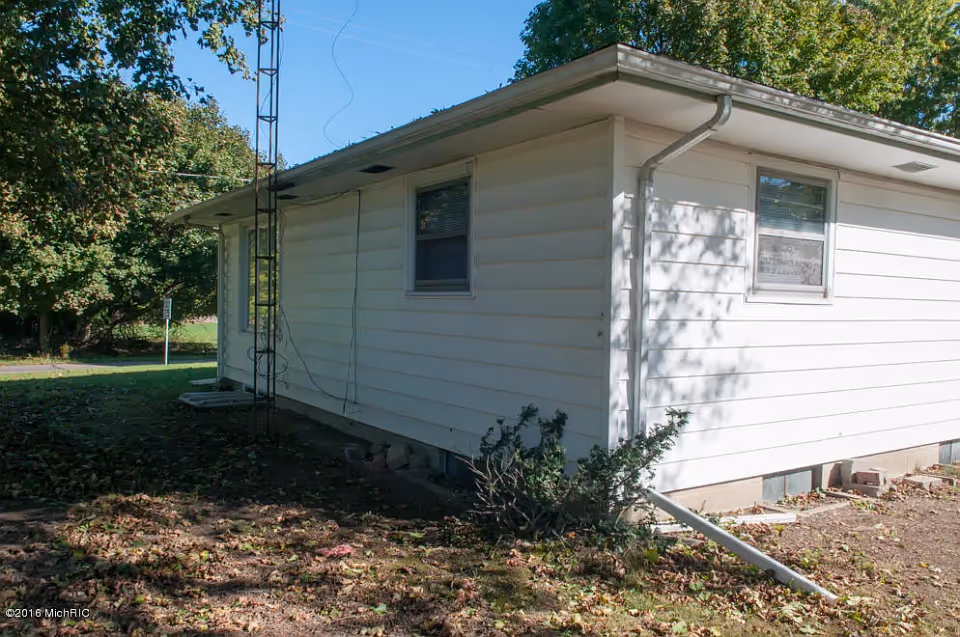 Exterior side view of a single-story white house with two windows, a metal antenna tower attached to the side, and some bushes and trees around the yard.