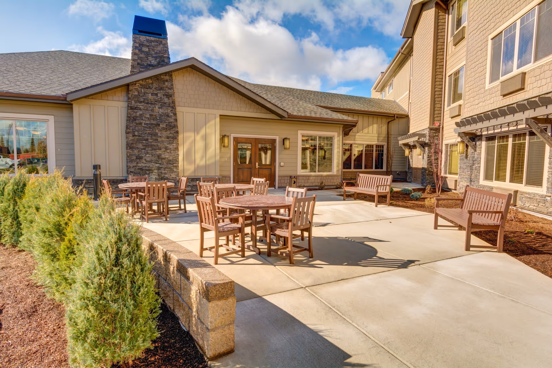 Sunlit outdoor patio with round wooden tables, chairs, and benches beside a multi-story assisted living building.
