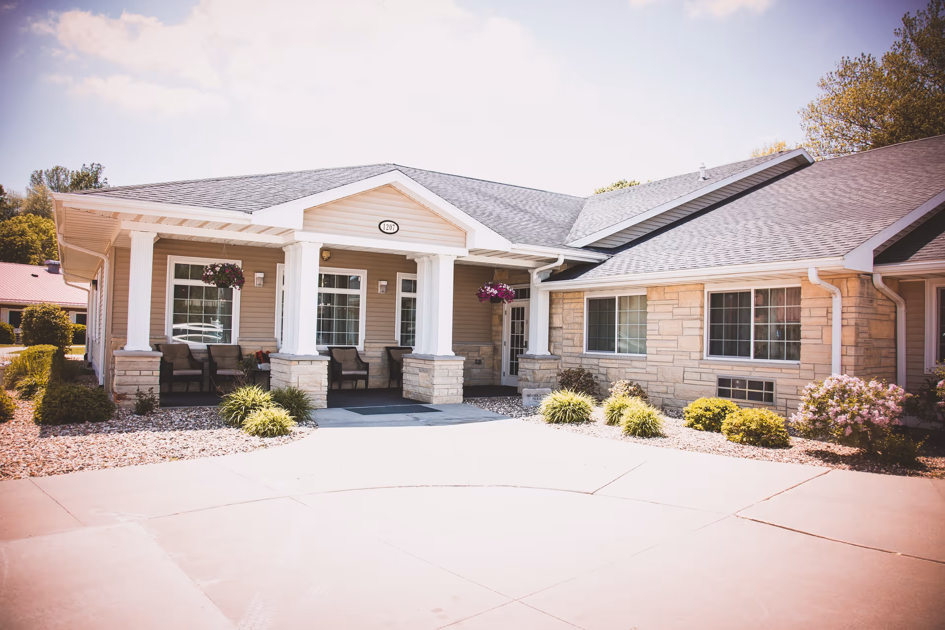 Exterior view of a single-story assisted living facility building with beige siding and stone accents. The entrance features a covered porch supported by white columns, with several chairs arranged on the porch. There are hanging flower baskets and landscaped bushes and plants around the building. The sky is partly cloudy with sunlight illuminating the scene.
