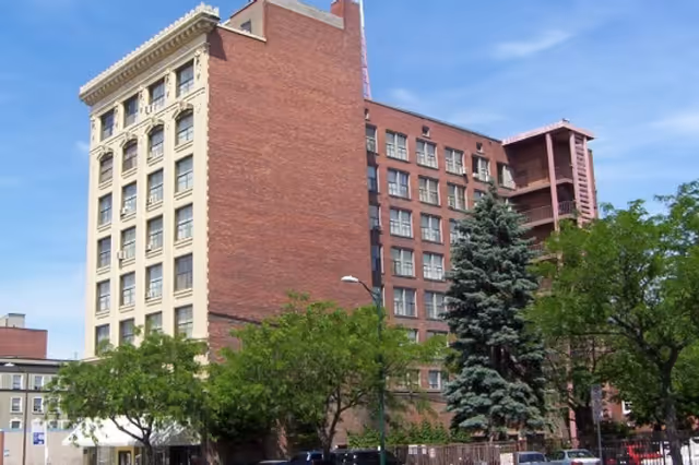 A tall brick apartment building with trees in the foreground and a clear blue sky.
