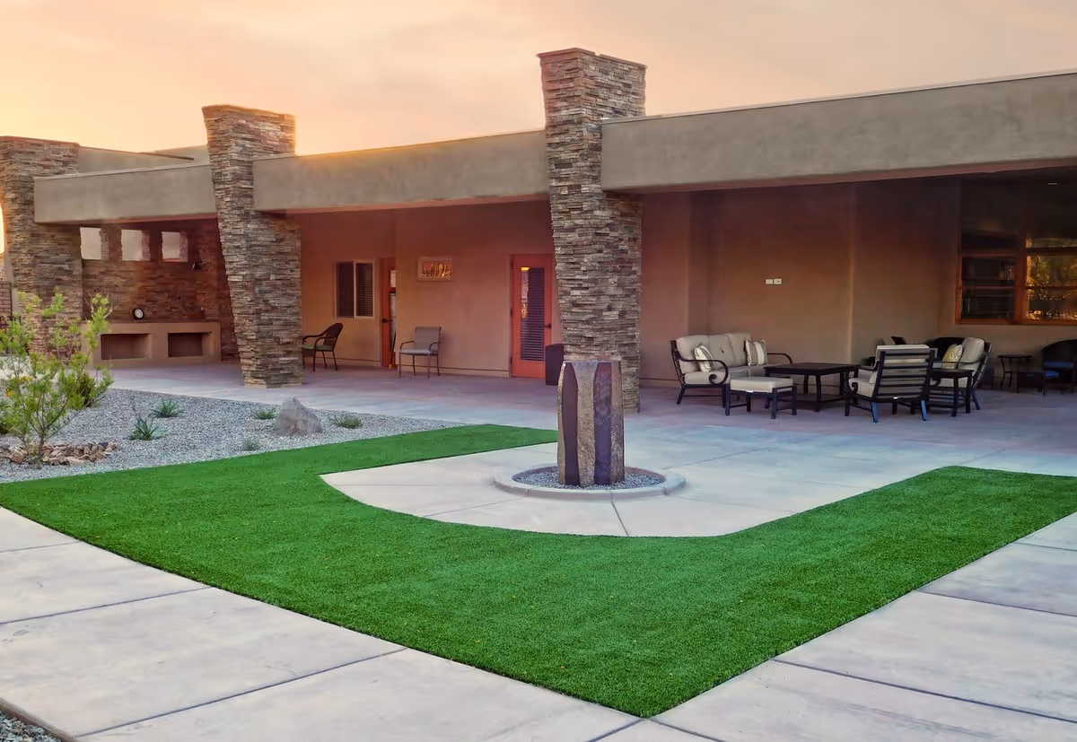 Outdoor patio area at Academy Villas featuring stone pillars, cushioned seating arrangements, a small green lawn, and a decorative stone sculpture in the center, with a sunset sky in the background.