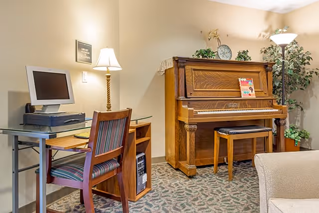 A cozy interior room featuring a wooden upright piano with a small bench, a computer desk with a monitor and printer, a wooden chair with striped upholstery, a floor lamp, a table lamp, and some decorative plants and items on top of the piano.