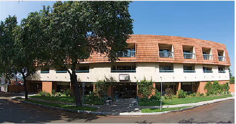 Exterior view of a three-story retirement residence building with a curved facade, featuring balconies on the upper floors, surrounded by trees and greenery under a clear blue sky.