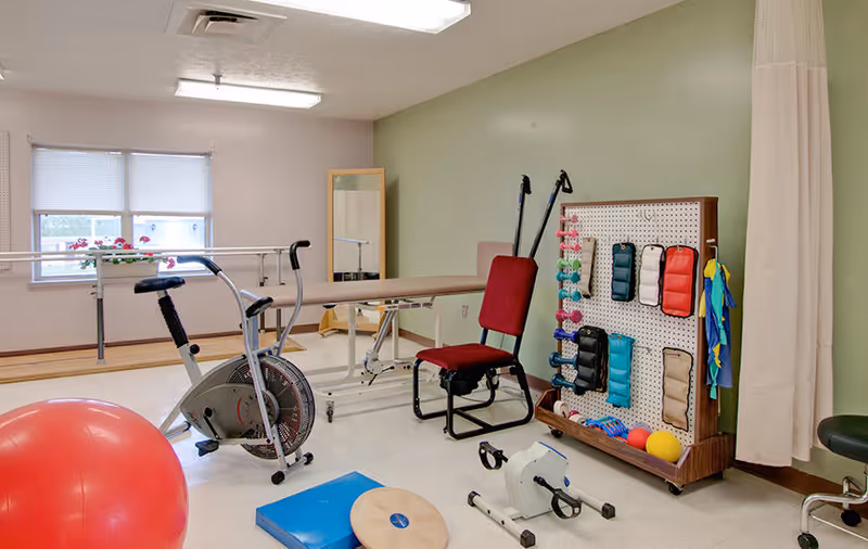 A physical therapy or exercise room with various equipment including a stationary exercise bike, a red exercise ball, a padded therapy table, a red chair, a pegboard with colorful weights and exercise bands, and a small pedal exerciser. The room has a green and beige wall, a window with blinds, and a full-length mirror.