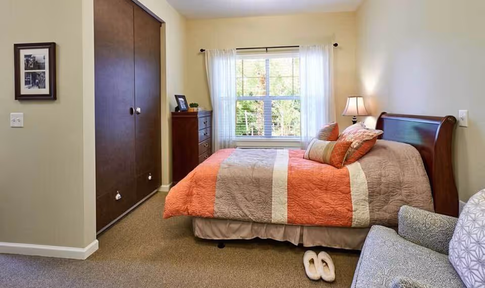 A cozy bedroom with a bed covered in a quilted orange and beige bedspread, several pillows, a wooden headboard, a nightstand with a lamp, a window with sheer white curtains, a wooden dresser, a framed picture on the wall, a pair of slippers on the carpeted floor, and a patterned armchair.