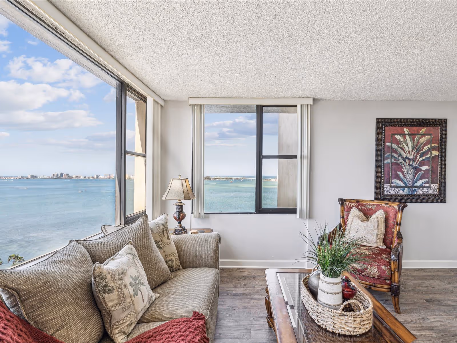 A bright living room with large windows offering a scenic view of a body of water and distant buildings. The room features a beige sofa with patterned cushions, a wooden coffee table with a woven basket and decorative items, a red upholstered armchair with a pillow, a side table with a lamp, and a framed botanical artwork on the wall.
