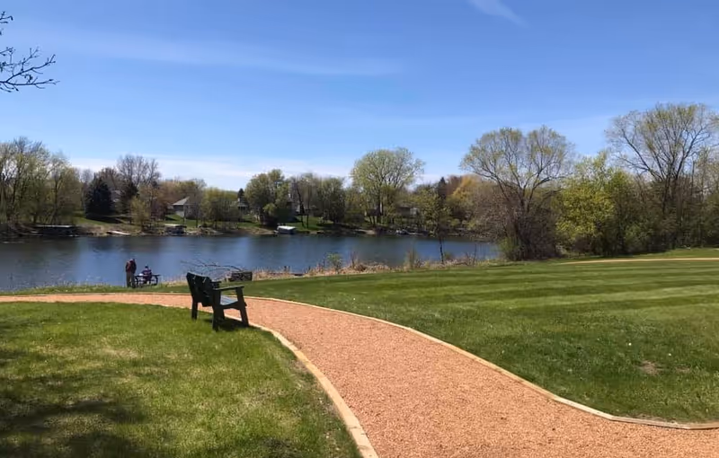 A curved gravel path with a bench beside a grassy lawn overlooks a calm lake with trees and two people near the shoreline under a blue sky.