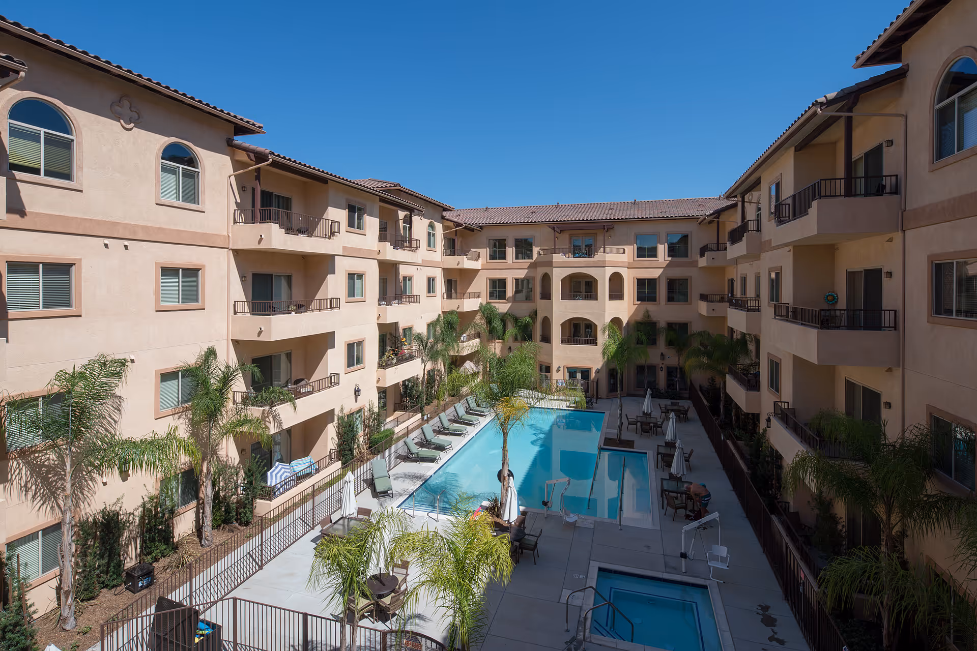 Courtyard-style apartment complex surrounding a rectangular swimming pool with lounge chairs and palm trees under a clear blue sky.
