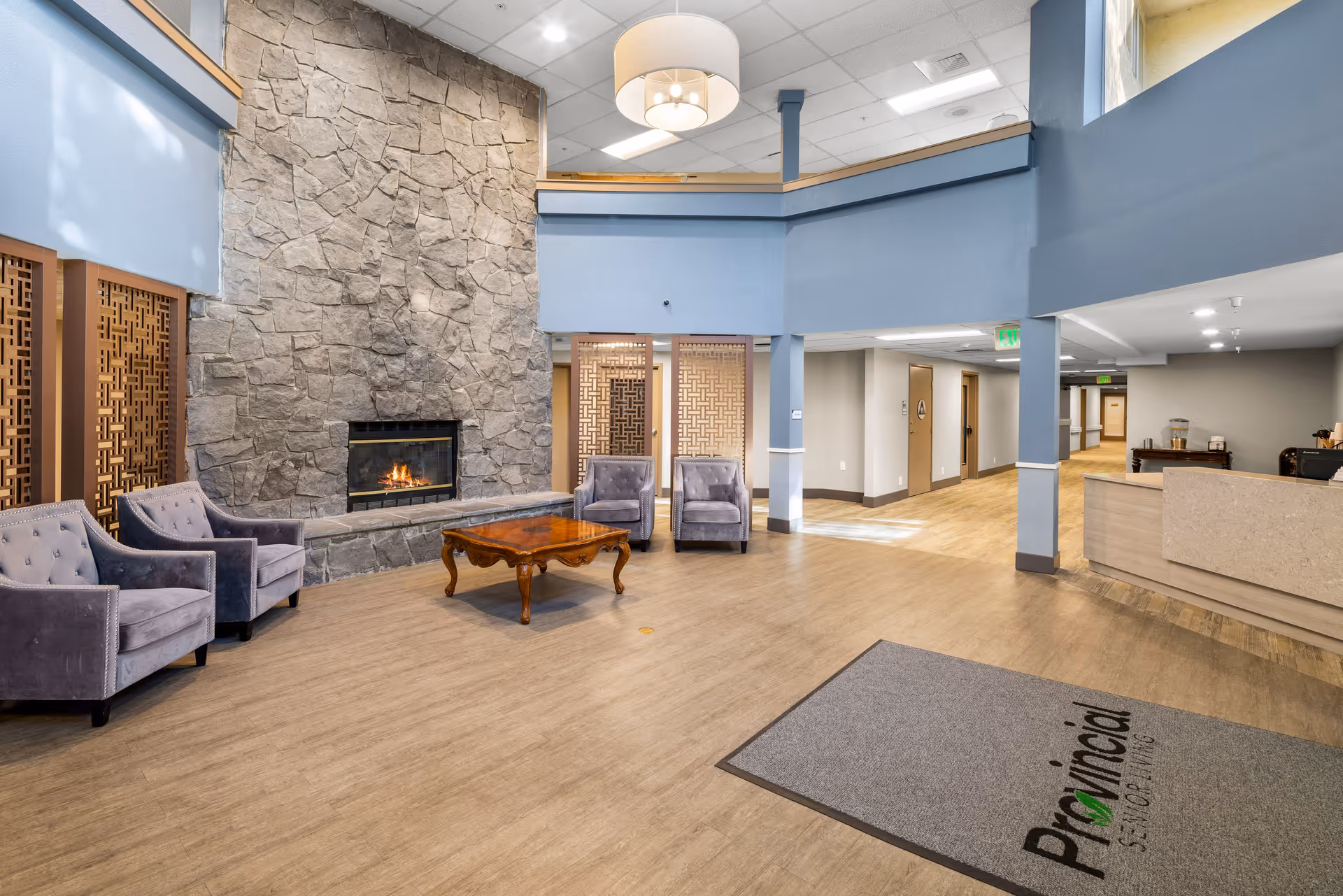 A spacious senior living community lounge area featuring a large stone fireplace with a fire burning, four gray upholstered armchairs arranged around a wooden coffee table, light wood flooring, blue walls with decorative wooden panels, and a reception desk to the right. A ceiling light fixture hangs above, and a gray floor mat with the text 'Provincial Senior Living' is visible near the entrance.