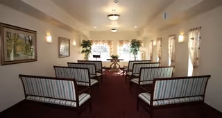 A small chapel or meeting room with rows of wooden benches featuring striped cushions, beige walls adorned with framed artwork, wall-mounted lights, and windows with sheer curtains letting in natural light. There are two potted plants near the front and a small table with a floral arrangement.