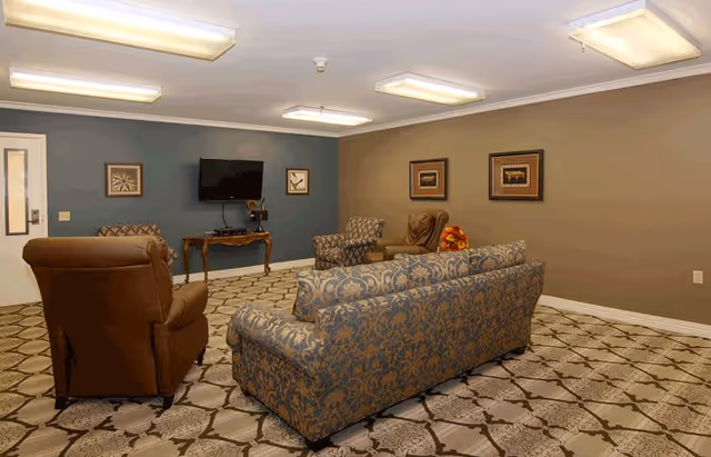 A cozy living room area with patterned carpet, a blue and gold patterned sofa, two armchairs, a small wooden table with a TV mounted on the wall above it, and framed artwork on the walls.