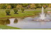 A small landscaped pond with a central fountain surrounded by grass, trimmed shrubs, and trees.