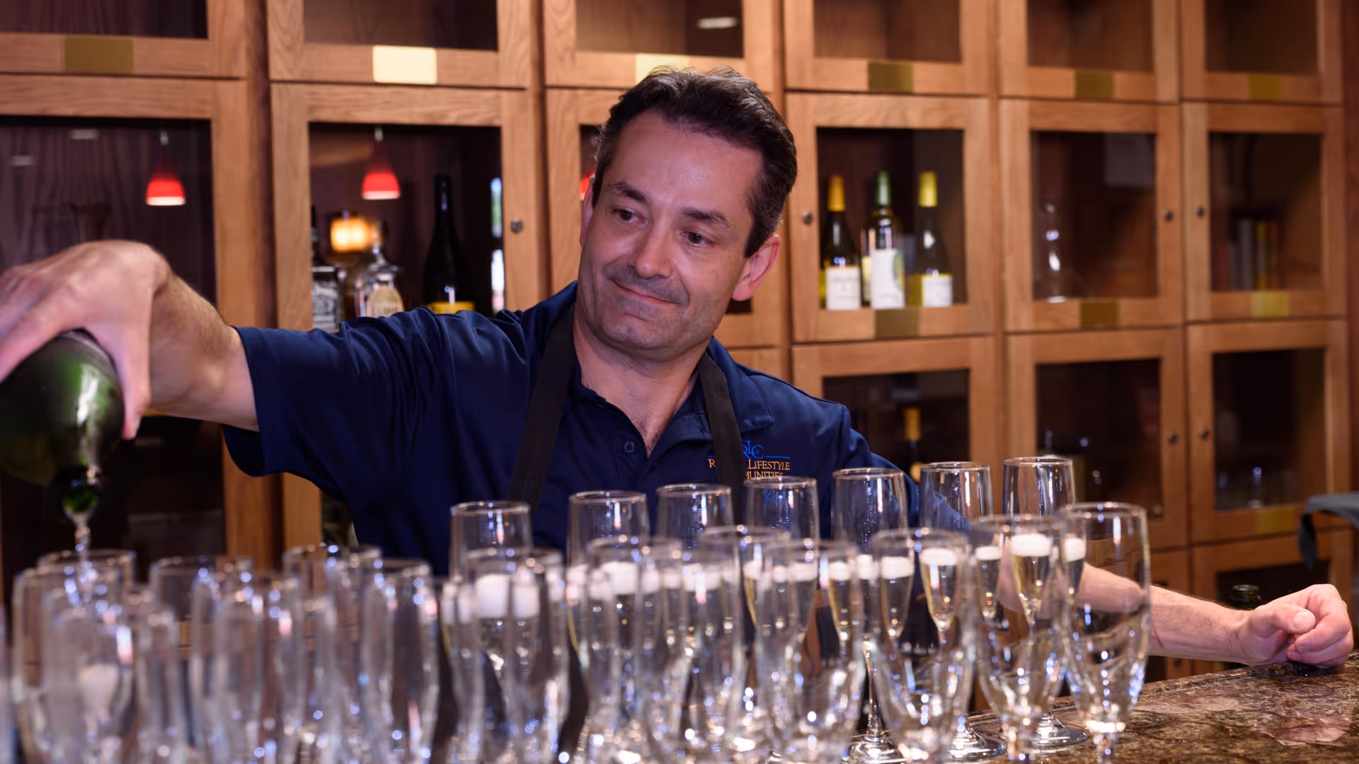A staff member behind a bar pours champagne into a row of flute glasses in front of wooden cabinets.