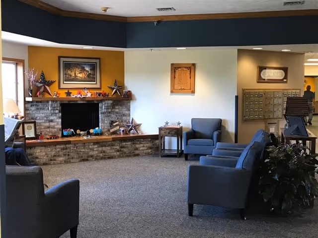 A cozy senior living facility common area with several blue upholstered armchairs arranged around a brick fireplace decorated with star ornaments and small decorative items. There is a framed picture above the fireplace and a plaque on the wall. Mailboxes are visible on the right side of the room, along with a potted plant and a small table.