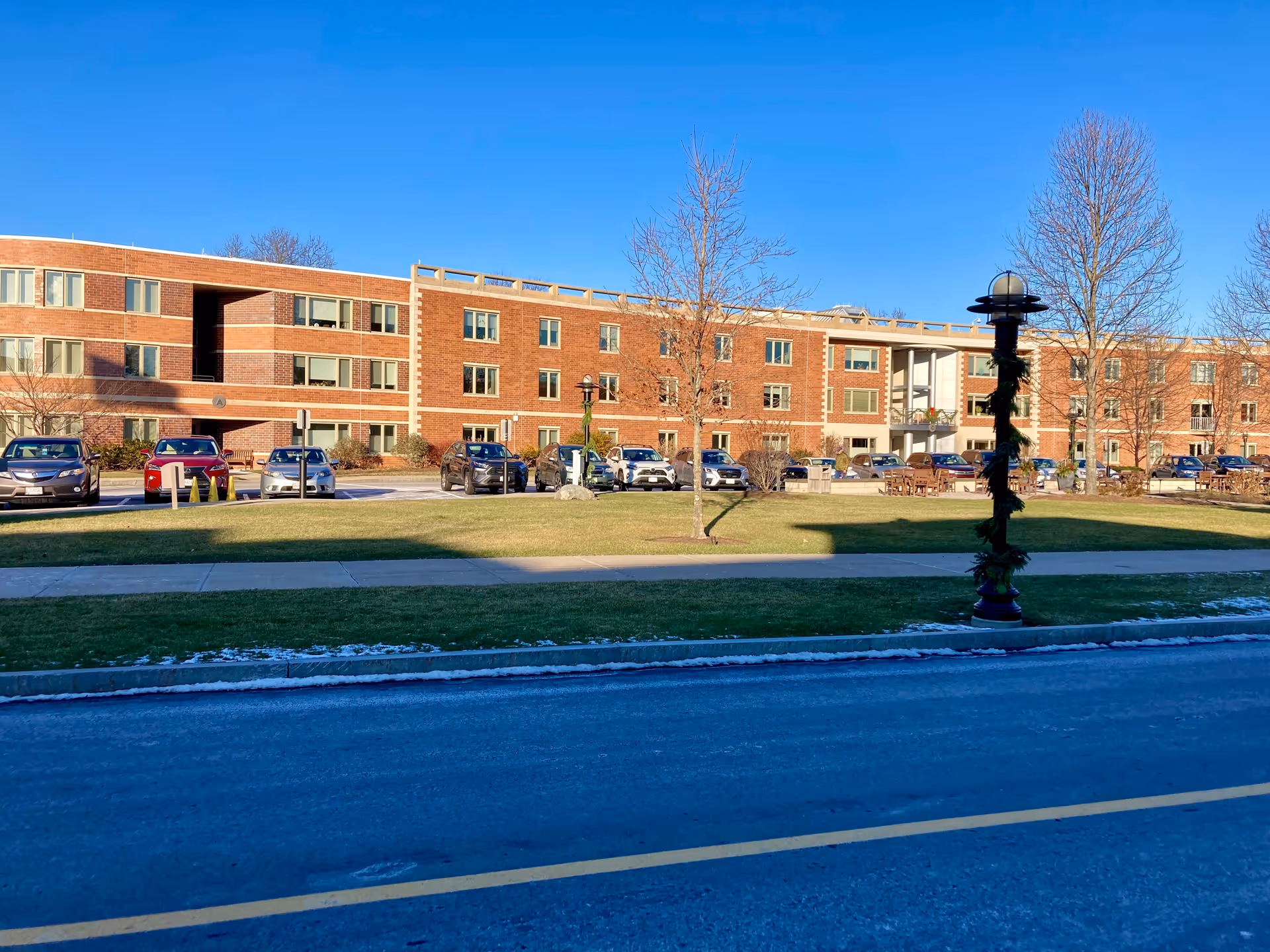Exterior view of a three-story brick building with multiple windows, a parking lot with several cars, a grassy area with leafless trees, and a streetlamp decorated with greenery under a clear blue sky.