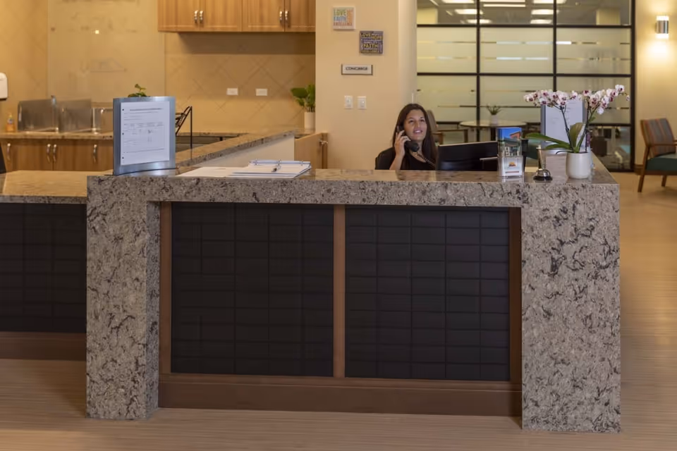 Reception desk area with a woman speaking on the phone behind the counter. The desk has a granite countertop with a potted orchid plant, a bell, and informational brochures. Behind the desk, there are cabinets, a sink, and a wall with a sign labeled 'CONCIERGE'. The background includes a glass partition and seating area.