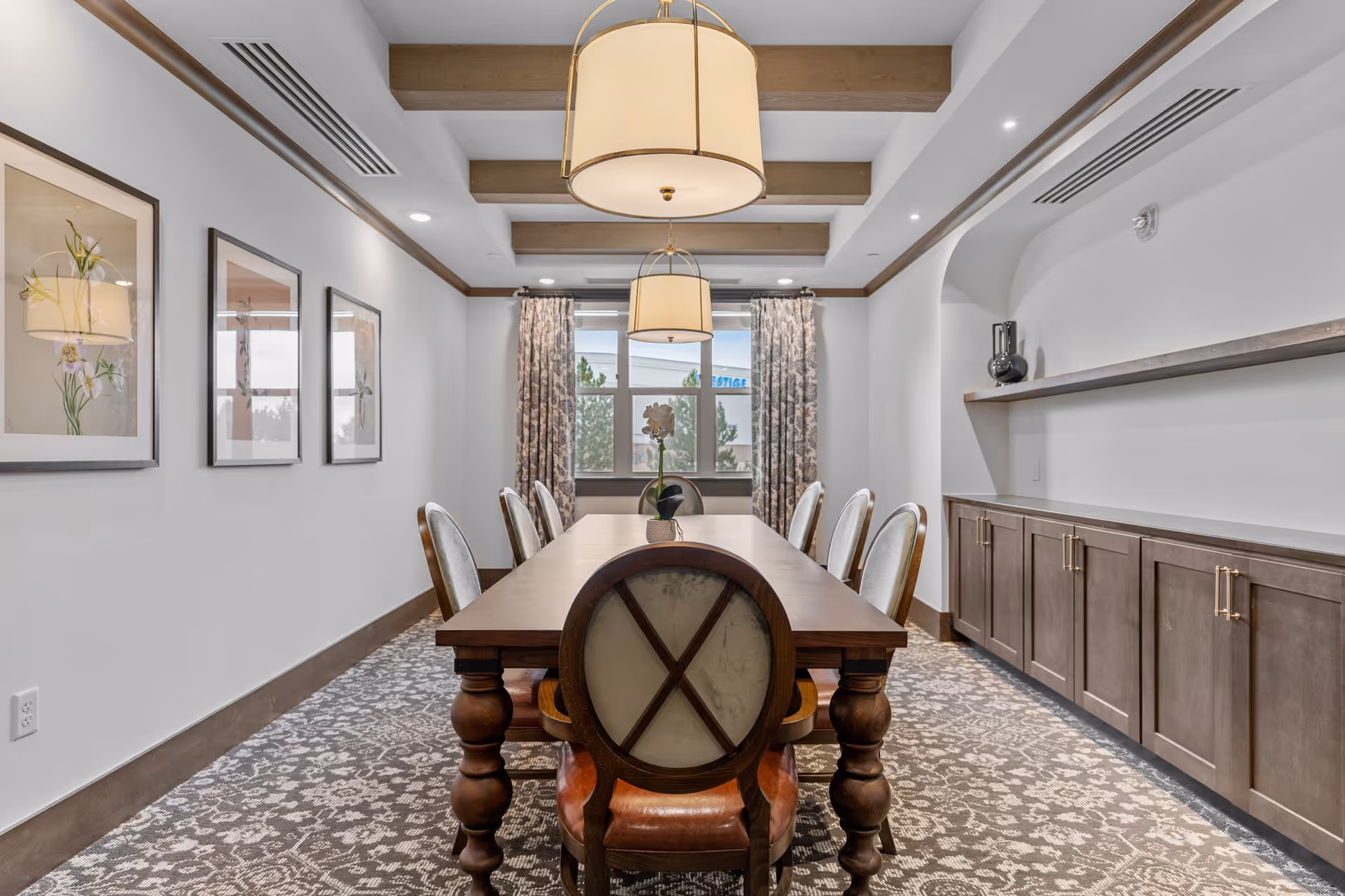 Long dining room with a wooden table and upholstered chairs, pendant lights, framed artwork, and built-in cabinets under a beamed ceiling.