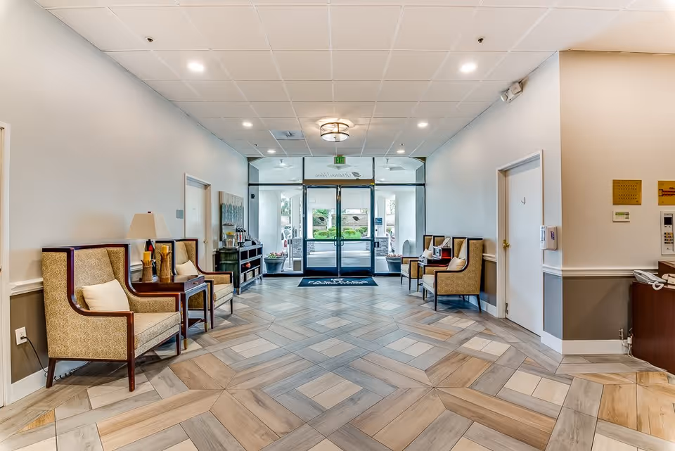 A spacious and well-lit senior living facility lobby with patterned tile flooring, beige walls, and a ceiling with recessed lighting. There are four upholstered armchairs with wooden frames and small tables with lamps and decorative items on either side of the room. At the far end, glass double doors lead outside, and a dark blue mat is placed in front of the doors. A small console table with a coffee station is positioned against the left wall.