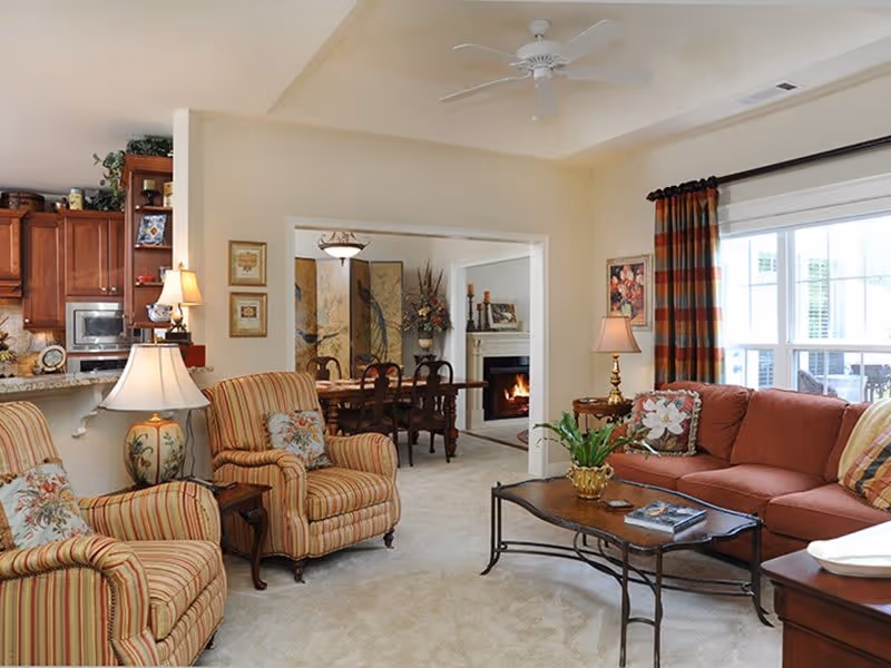 A cozy living room with a red sofa, two striped armchairs with floral pillows, a wooden coffee table with a plant and books, and two table lamps. The room opens into a dining area with a wooden dining table and chairs, a fireplace, and decorative artwork on the walls. The kitchen with wooden cabinets and a microwave is partially visible to the left.