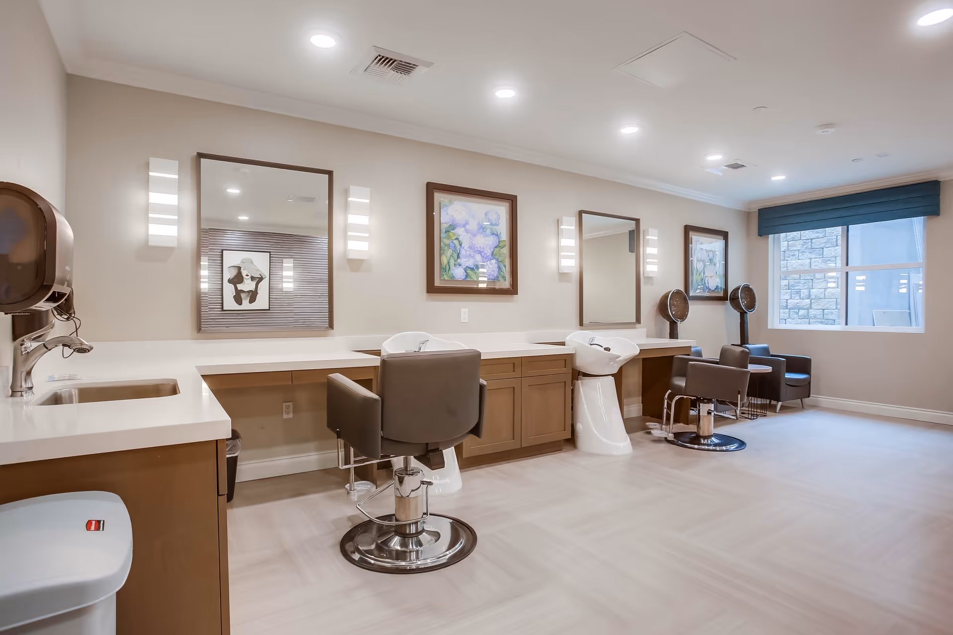 A bright and clean salon area in a senior living facility with two styling chairs in front of large mirrors, two hair washing stations, two hair dryers, and framed artwork on the walls. The room has light-colored flooring, recessed ceiling lights, and a window with a blue valance.