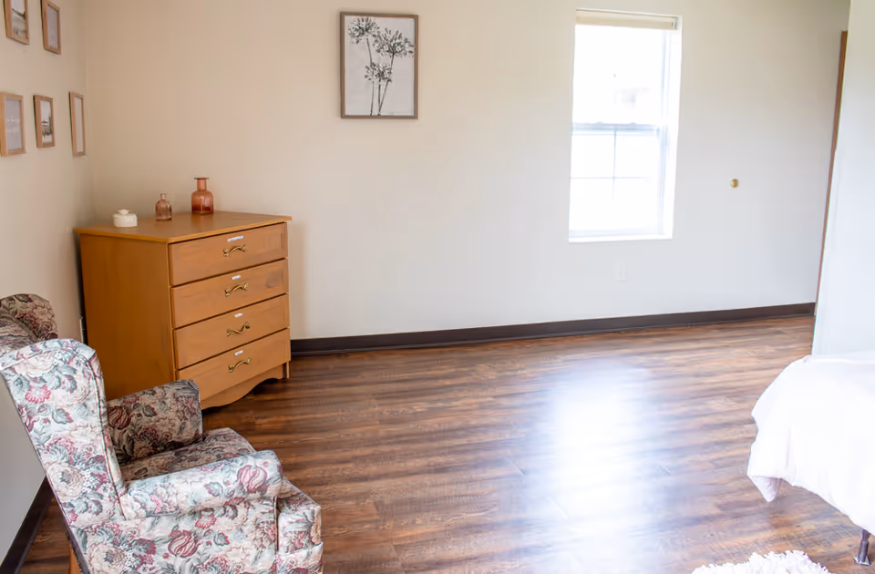 Sparsely furnished bedroom with a floral armchair, wooden dresser, window, and part of a bed on wood floors.