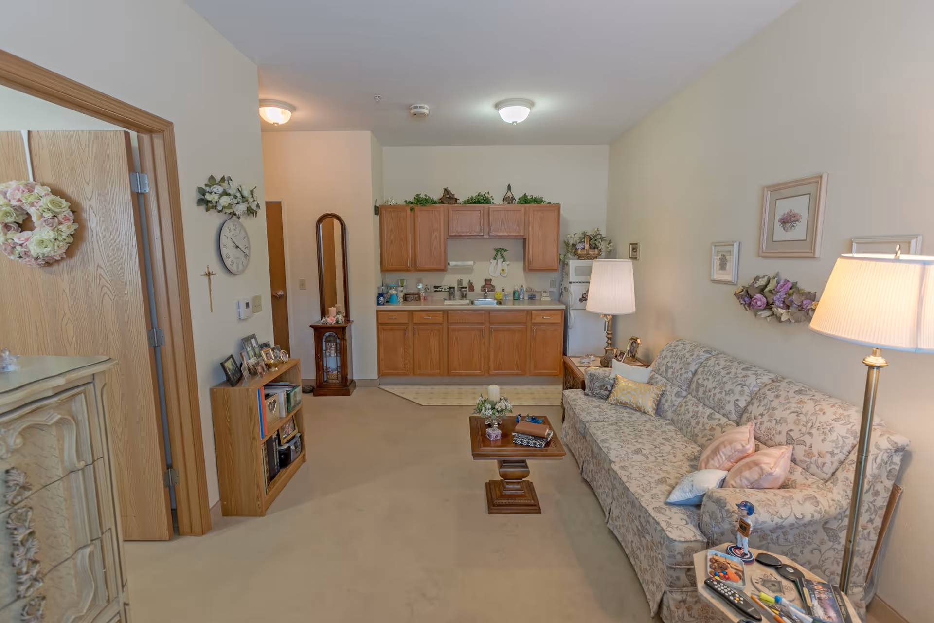 A cozy living area in a senior living facility featuring a floral-patterned sofa with multiple pillows, a wooden coffee table with decorative items, and two standing lamps providing warm lighting. The room includes a small kitchen area with wooden cabinets, a sink, and a refrigerator. The walls are decorated with framed floral artwork and a floral wreath. There is a wooden bookshelf with picture frames and a clock on the wall near a doorway.