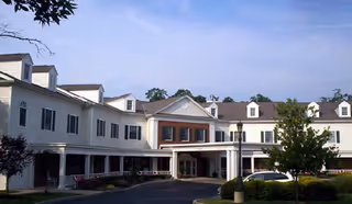Front exterior of a white multi-story senior living building with a covered entrance, columns, and surrounding landscaping.