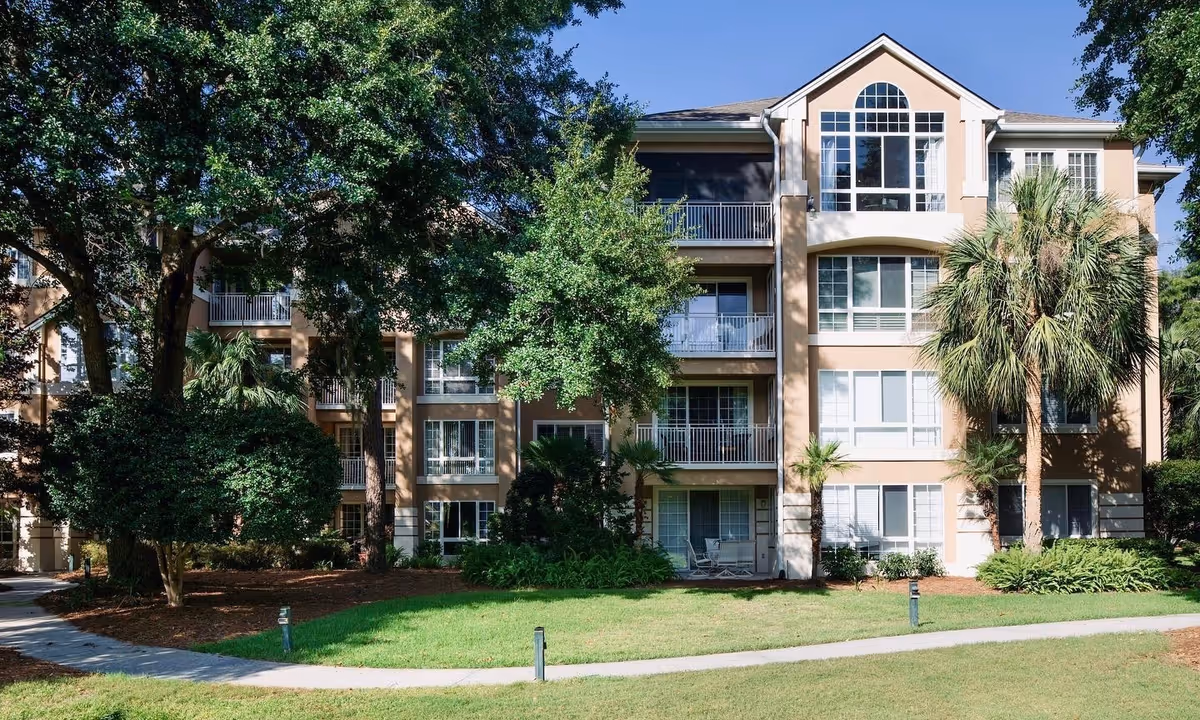 Front exterior of a multi-story senior living building with balconies, palm trees, and a grassy lawn.